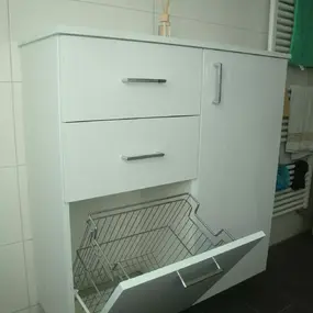 Weißer Badezimmerschrank mit Schubladen und ausklappbarem Wäschekorb auf dunklem Boden.